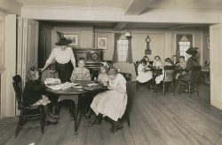 Caroline Emmerton is standing talking to children as they write around a table in the House of the Seven Gables.