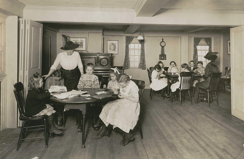 Caroline Emmerton is standing talking to children as they write around a table in the House of the Seven Gables.
