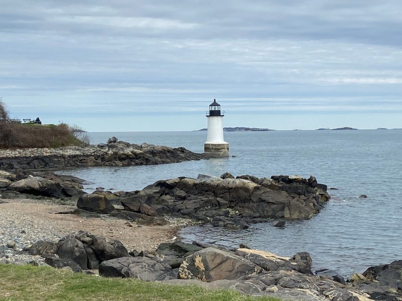View of the Winter Island light house with a rocky beach in the foreground and the Atlantic Ocean in the background on a slightly cloudy day.