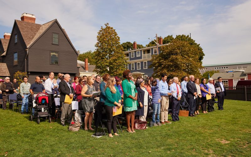 A large group of people stand in rows on the Gables' lawn to take their oath of allegiance for United States citizenship.