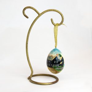 The front of the House of the House of the Seven Gables Painted Egg Ornament. It is hanging from a gold stand.