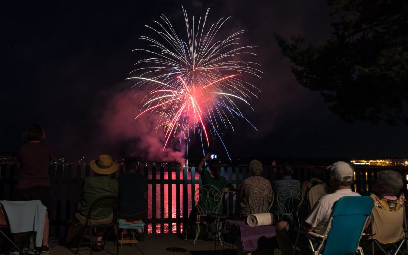 Multi-colored fireworks explode in the air over Salem Harbor as people in the foreground, facing away from the camera, watch.