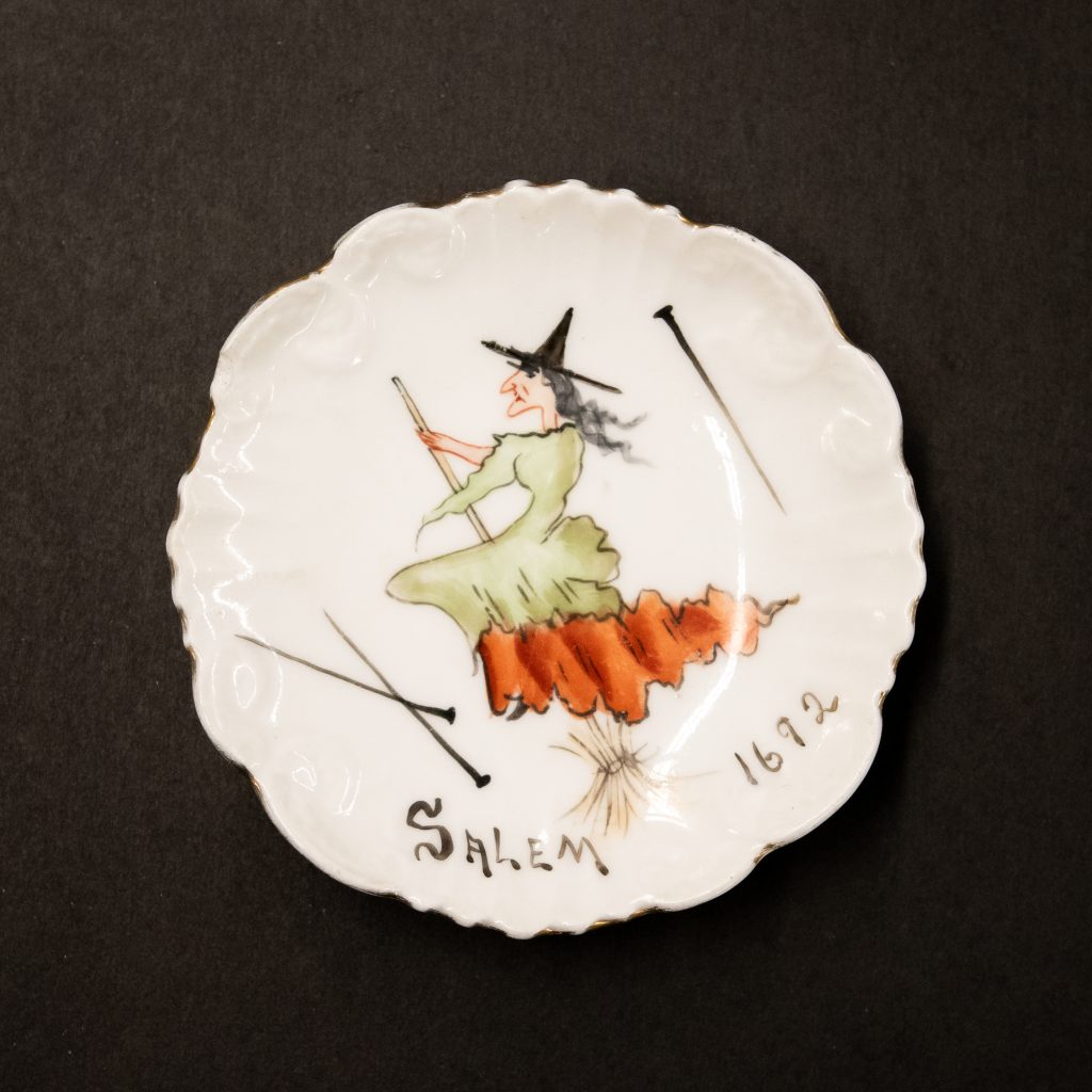 Painted ceramic dish with an image of a witch flying on a broomstick. Above the witch are pins, as some of the girls who were accusers claimed that a witch had pricked them with a pin. Below the witch is “Salem 1692” hand-painted in black paint.